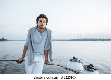 A Stylish Guy Stands At The Stern Of The Yacht, Resting On The Yacht At Sunset, A Pensive Look. He Wears White Shorts, Gray Sweatshirt On Her Shoulders. Summer Cruise