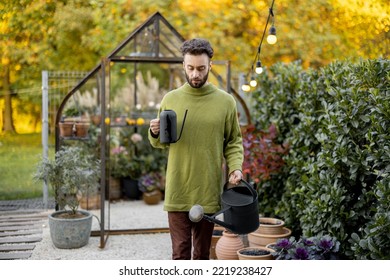 Stylish Guy Watering Plants, Taking Care Of Them In Beautiful Garden With Small Glass House On Background. Hobby, Work In Garden Concept