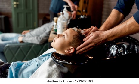 Stylish haircut for child. Side view of a cute boy leaning on the sink in barbershop while male barber washing his hair. Child in hairdressing salon. Children's hairdresser. Kids in barbershop