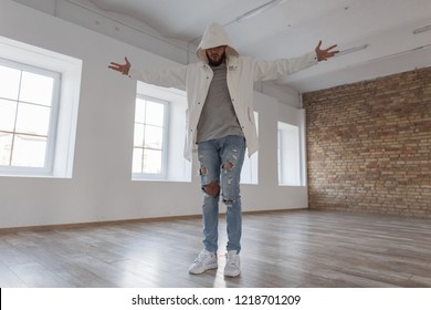Stylish handsome attractive model man in a jacket with a hood in fashion torn jeans with white sneakers posing in a white room with a brick wall