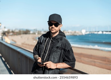 Stylish handsome fashion hipster man with sunglasses with cap in black denim jacket with hoodie holding smartphone and walking along beach in Barcelona, ​​Spain - Powered by Shutterstock