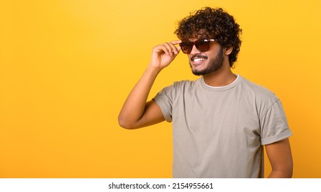 Stylish handsome Indian young curly man wearing casual t-shirt and sunglasses has good mood, enjoying sunny day, posing isolated on yellow background - Powered by Shutterstock