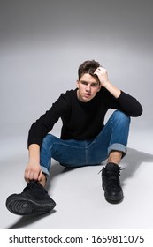 Stylish handsome man posing sitting on floor. Handsome Guy. cool fashion male model sitting on grey background and looking at the camera. Student in a studio in jeans and a black sweater.