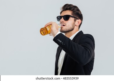 Stylish Handsome Man In Sunglasses And Black Suit Drinking From Beer Bottle Over Grey Background