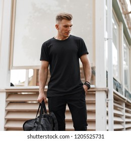 Stylish handsome young american hipster model man with hairstyle in black t-shirt with fashion bag standing on the street near the wooden building