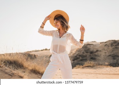 stylish happy attractive smiling woman posing in desert sand dressed in white clothes wearing straw hat and sunglasses on sunset, travel safari on vacation, sunny summer day