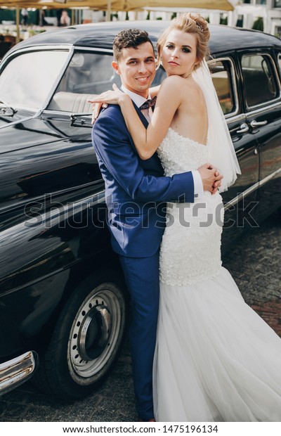 Stylish happy bride and groom embracing\
at old black retro car. Gorgeous wedding couple of newlyweds\
hugging after wedding ceremony. Romantic\
moment