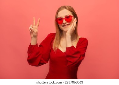 Stylish happy smiling young woman in red dress, french beret and trendy heart shaped glasses showing peace gesture v sign with two fingers and keeping hand on face isolated on colored pink background.