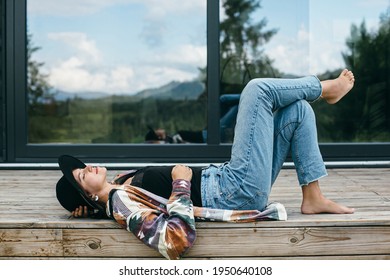 Stylish happy woman relaxing on wooden terrace on background of modern cabin with windows. Young female in casual cloth and hat lying on porch in mountains. Calm tranquil moment. Travel