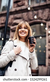 Stylish happy young woman wearing beige trench coat.She holds coffee to go. portrait of smiling girl . Street fashion concept.