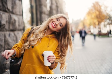 Stylish happy young woman wearing boyfriend jeans, white sneakers bright yellow sweatshirt.She holds coffee to go. portrait of smiling girl in sunglasses and bag