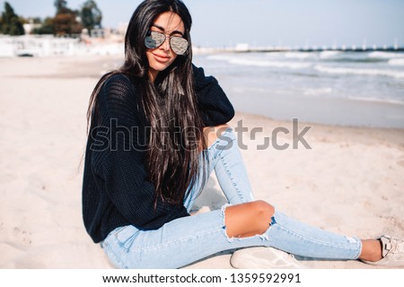 Stylish happy young woman wearing boyfriend jeans, black sweater and white sneakers sitting on the beach.Lifestyle photo of weekend time.woman in fashionable ripped Jeans 
