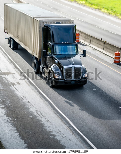 Stylish Heavy loaded classic black big rig semi\
truck with roof spoiler transporting commercial cargo at container\
on semi trailer running on the straight wide divided multiline\
highway road