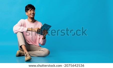 Stylish hindu guy with digital tablet sitting on floor and looking at copy space, checking newest mobile application, trading online or chating with ladies on dating app, panorama, blue background