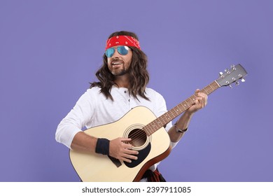 Stylish hippie man in sunglasses playing guitar on violet background