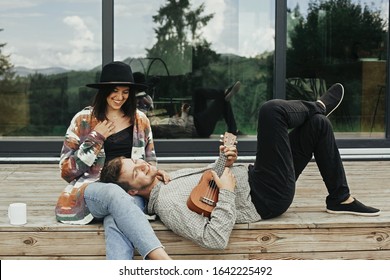 Stylish hipster couple sitting on wooden porch, relaxing with ukulele  on background of modern cabin with big windows in mountains. Happy young family travelers enjoying vacation in woods - Powered by Shutterstock