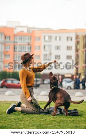 Stylish hipster girl dresses the dog on the lawn against the background of urban landscape, looks at the dog and smiles. Happy lady playing with dog on a walk. Vertical portrait of owner and dog