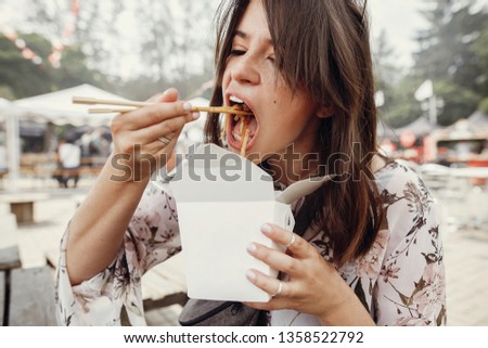 Stylish hipster girl eating wok noodles with vegetables from carton box with bamboo chopsticks. Asian Street food festival. Boho woman eating thai noodles in takeaway paper box. Food delivery