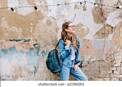 Stylish hipster girl in the retro jeans suit posing in front of the old brick wall. Trendy young woman with bag standing next to old building. Street fashion concept, wearing glasses