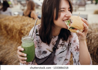 Stylish Hipster Girl In Sunglasses Eating Delicious Vegan Burger And Holding Smoothie In Glass Jar In Hands At Street Food Festival. Happy Boho Woman Biting Burger With Drink In Summer Street