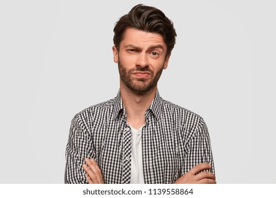 Stylish hipster male keeps arms folded, looks with hesitant expression, raises eyebrow with bewilderment, listens somebody`s suggestion, doubts whether to agree or not. Facial expressions concept