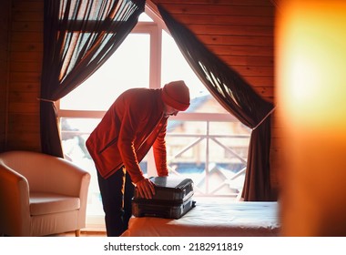 Stylish Hipster Traveller Man In Red Hat In Wooden House Packing Suitcase, Winter Travel Concept, Space For Text, Comfy Moment. Holiday And Travel Concept