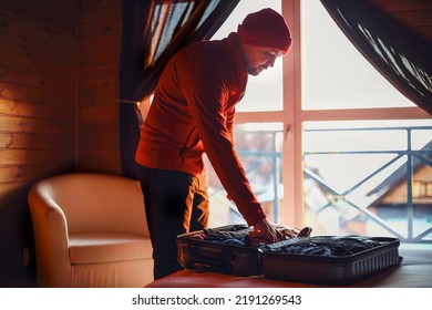 Stylish Hipster Traveller Man In Red Hat In Wooden House Packing Suitcase, Winter Travel Concept, Space For Text, Comfy Moment. Holiday And Travel Concept