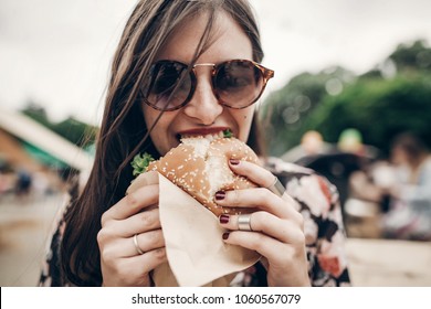 Stylish Hipster Woman Eating Juicy Burger. Boho Girl Biting Yummy Cheeseburger, Smiling At Street Food Festival. Summertime. Summer Vacation Travel Picnic. Space For Text