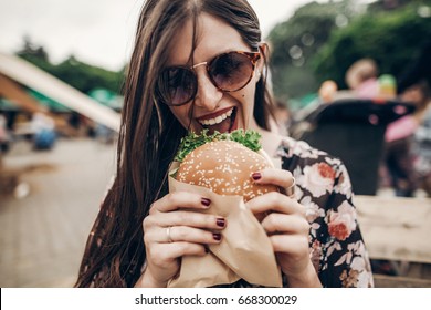 Stylish Hipster Woman Eating Juicy Burger. Boho Girl Biting Cheeseburger, Smiling At Street Food Festival. Summertime. Summer Vacation Travel Picnic. Space For Text