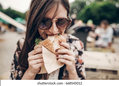 Stylish Hipster Woman Eating Juicy Burger. Boho Girl Biting Yummy Cheeseburger, Smiling At Street Food Festival. Summertime. Summer Vacation Travel Picnic. Space For Text