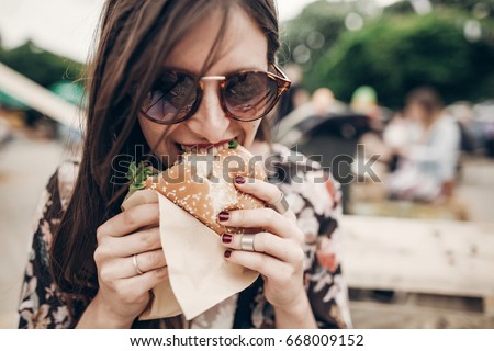 stylish hipster woman holding juicy burger and eating. boho girl biting hamburger  smiling at street food festival. summertime. summer vacation travel. space for text
