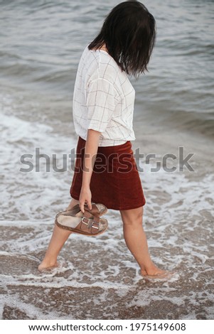 Stylish hipster woman walking barefoot on sandy beach with sea waves in the evening, enjoying calm moment. Casual young female with flip flops in hand relaxing on seashore