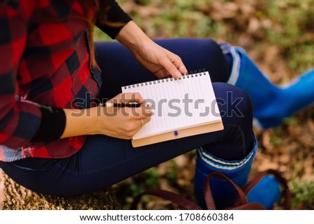 stylish hipster young woman sitting on a tree trunk, with a backpack, thermo mug and notebook in the forest on sunset light in the spring season, looking at amazing woods, travel concept