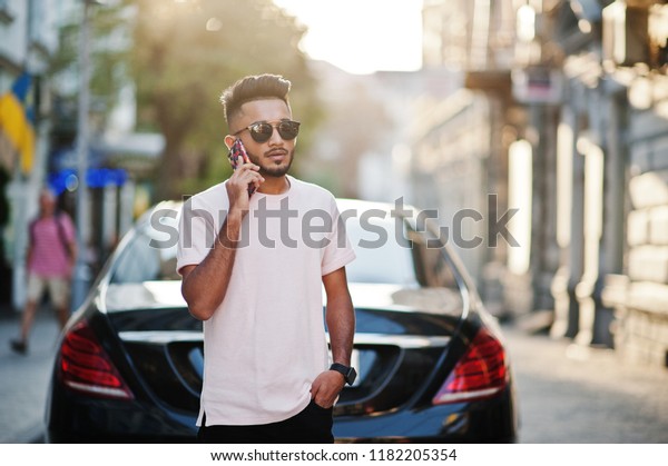 Stylish indian beard man at sunglasses\
and pink t-shirt against luxury car and speaking on mobile phone.\
India rich model posed outdoor at streets of\
city.