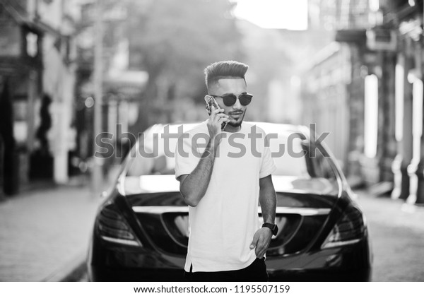 Stylish indian beard man at sunglasses\
and pink t-shirt against luxury car and speaking on mobile phone.\
India rich model posed outdoor at streets of\
city.