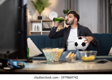 Stylish Indian Guy Drinking Cold Beer While Sitting Comfortably On Couch And Enjoying Football Championship At Home. Concept Of Free Time, Sport And Entertainment.