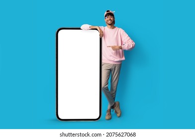 Stylish indian guy standing by huge smartphone with blank screen, mockup, pointing at advertisement, smiling millennial man recommending newest mobile application, blue studio background, copy space - Shutterstock ID 2075627068