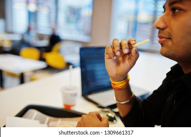 Stylish Indian Man Sitting At Fast Food Cafe Against His Laptop And Eating French Fries.