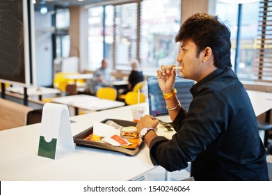 Stylish Indian Man Sitting At Fast Food Cafe Against His Laptop And Eating French Fries.