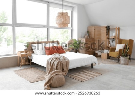 Stylish interior of light bedroom with wooden chest of drawers, folding screen, armchair and bed