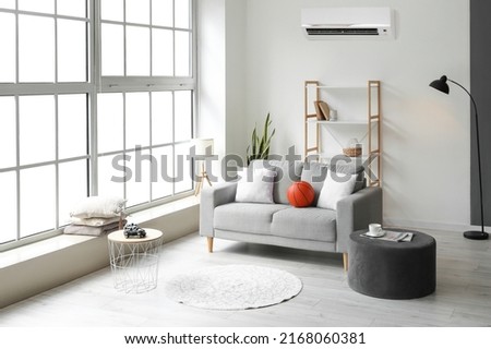 Stylish interior of light living room with sofa, shelf unit, pouf and air conditioner