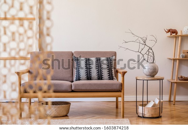 Stylish interior of living room at cozy
apartment with brown wooden sofa, coffe table, bookstand , pillow,
flowers and elegant accessories. Beige and japandi concept. Modern
home staging. Template.