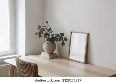 Stylish interior. Living room with poster frame mock up on wooden table, desk. Eucalyptus tree branches, rattan chair near window. Minimalist concept of home decor. Scandinavian design. Blank template - Powered by Shutterstock