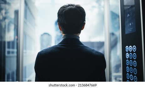 Stylish Japanese Businessman Riding Glass Elevator To Office In Modern Business Center. Male Looking At Modern Downtown Skyscrapers Out Of The Panorama Window In The Lift. Back Turned To Camera