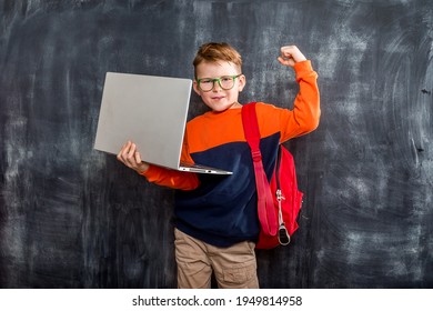 Stylish Kid Boy Ready For School. 7-8 Years Old Student With Backpack Using Laptop. Child In Orange Shirt And Red Backpack Against Blackboard Background With Free Space.