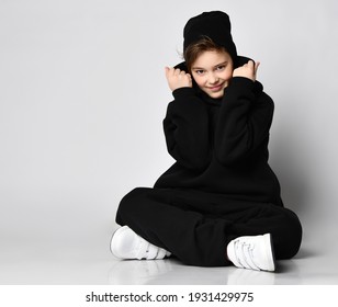 Stylish kid bro sits on in a lotus against a white wall and adjusts his hood. He is wearing a trendy black hoodie and sweatpants. black beanie and white sneakers