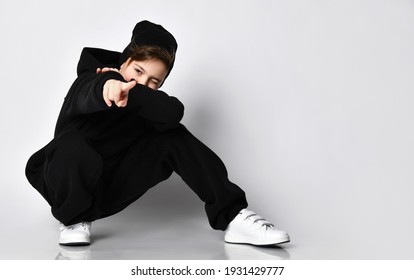 Stylish kid bro is squatting against a white wall and pointing at you. He is wearing a trendy black hoodie and sweatpants. black beanie and white sneakers - Powered by Shutterstock