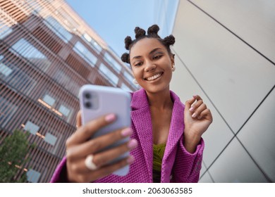Stylish lady with hairstyle dressed in fashionable pink jacket takes selfie on mobile phone for social networks poses against blurred city buildings smiles happily enjoys free time streams video