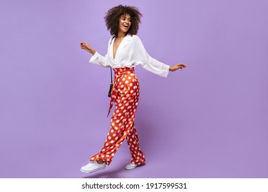 Stylish lady in wide pants, sneakers and blouse posing on isolated backdrop. Trendy woman with wavy hair smiling on lilac background..