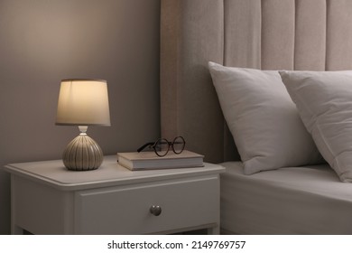 Stylish lamp, book and glasses on bedside table indoors. Bedroom interior elements